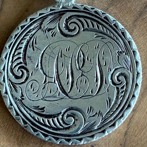 May include: A circular silver pendant featuring an intricate engraved monogram of the letter W, surrounded by decorative scrollwork and a textured border. The pendant hangs from a silver rolo chain, showcasing a polished metallic finish.
