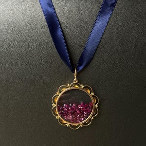 May include: A circular gold pendant featuring a clear glass center filled with loose, faceted deep pink rubies. The pendant has a scalloped, decorative gold frame and is suspended from a dark navy blue satin ribbon necklace.