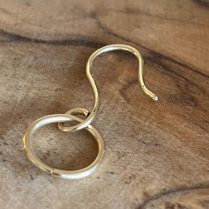 Könnte beinhalten: Ein goldfarbenes Ohrringelement aus Metall, bestehend aus einem gebogenen Haken, der an einem kreisförmigen Ring befestigt ist. Der Ohrhaken hat eine glatte, polierte Oberfläche und ein einfaches, funktionales Design für die Schmuckmontage.