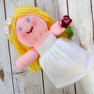 May include: A knitted doll wearing a white dress and holding a bouquet of pink and red flowers. The doll has blonde yarn hair and a pink face with blue eyes.
