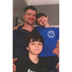 May include: A family portrait featuring a man and two children. The man wears a dark blue top. One child wears a blue t-shirt with a circular emblem, and the other wears a black t-shirt. All three are smiling.