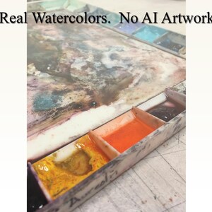 May include: A close-up of a watercolor paint palette with four wells. The wells contain orange, yellow, and dark purple paint. The palette is made of white plastic and has a worn, used appearance. The text "Real Watercolors. No AI Artwork" is visible at the top of the image.