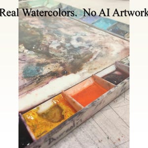 May include: A close-up of a watercolor paint palette with various colors, including yellow, orange, and dark purple. The text "Real Watercolors. No AI Artwork" is visible at the top of the image. The palette is made of a light-colored material.