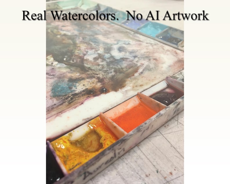 May include: A close-up of a watercolor paint palette with various colors, including yellow, orange, and dark purple. The text "Real Watercolors. No AI Artwork" is visible at the top of the image. The palette is made of a light-colored material.