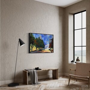 May include: A framed watercolor painting depicts a tree-lined street leading to a white house under a blue sky. The artwork is displayed on a neutral-toned wall above a wooden bench. A black floor lamp and a woven chair are also in the room.