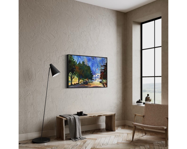May include: A framed watercolor painting depicts a tree-lined street leading to a white house under a blue sky. The artwork is displayed on a neutral-toned wall above a wooden bench. A black floor lamp and a woven chair are also in the room.