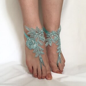 May include: A pair of light blue barefoot sandals featuring intricate floral and leaf lace embroidery. The design includes a central rose motif with delicate vine patterns that extend across the top of the foot, secured by a thin elastic strap.