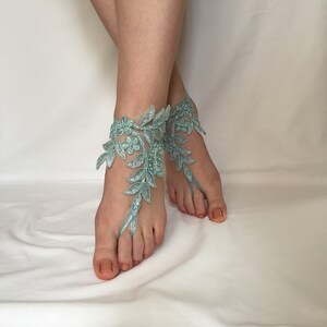 May include: A pair of light blue lace barefoot sandals featuring intricate floral and leaf embroidery. The design wraps around the ankle and includes a loop that secures around the second toe, embellished with small beads and metallic thread accents.