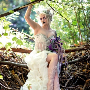 May include: A strapless white corset dress with a sweetheart neckline and crystal embellishments, paired with intricate floral lace leg wraps. The garment features a layered, flowing skirt and is worn with a headpiece featuring curved horns.