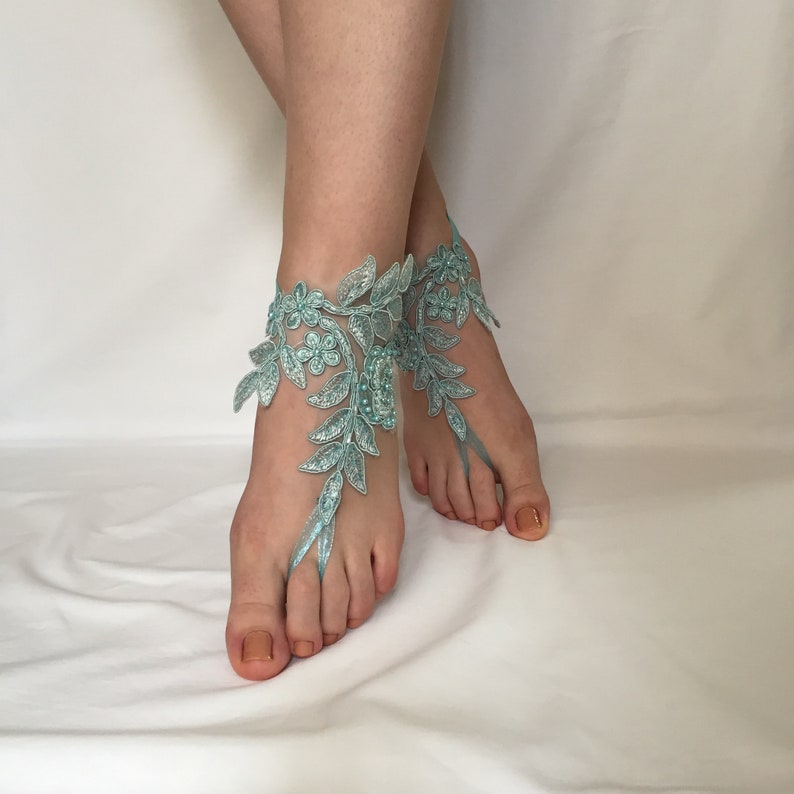 May include: A pair of light blue lace barefoot sandals featuring intricate floral and leaf embroidery. The design wraps around the ankle and includes a loop that secures around the second toe, embellished with small beads and metallic thread accents.
