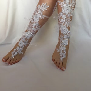 May include: A pair of white lace barefoot sandals featuring intricate floral embroidery, clear sequins, and small white beads. The design extends from a toe loop up the foot and along the lower leg, secured with thin white ribbons tied at the back.