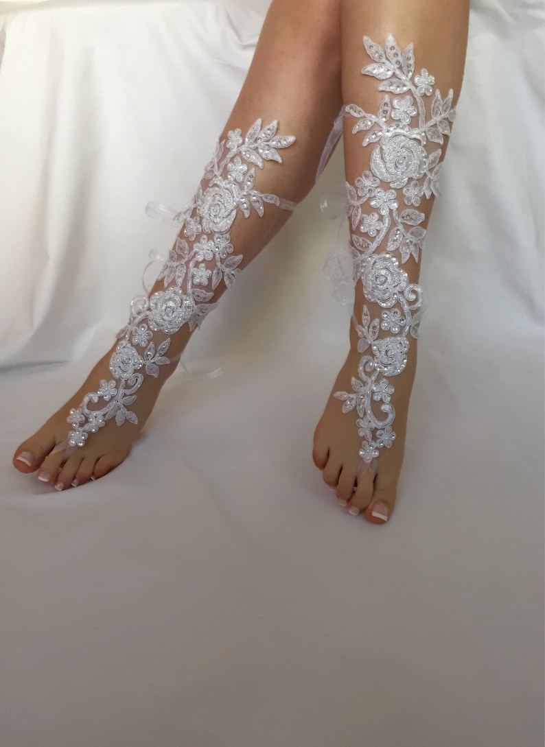 May include: A pair of white lace barefoot sandals featuring intricate floral embroidery, clear sequins, and small white beads. The design extends from a toe loop up the foot and along the lower leg, secured with thin white ribbons tied at the back.