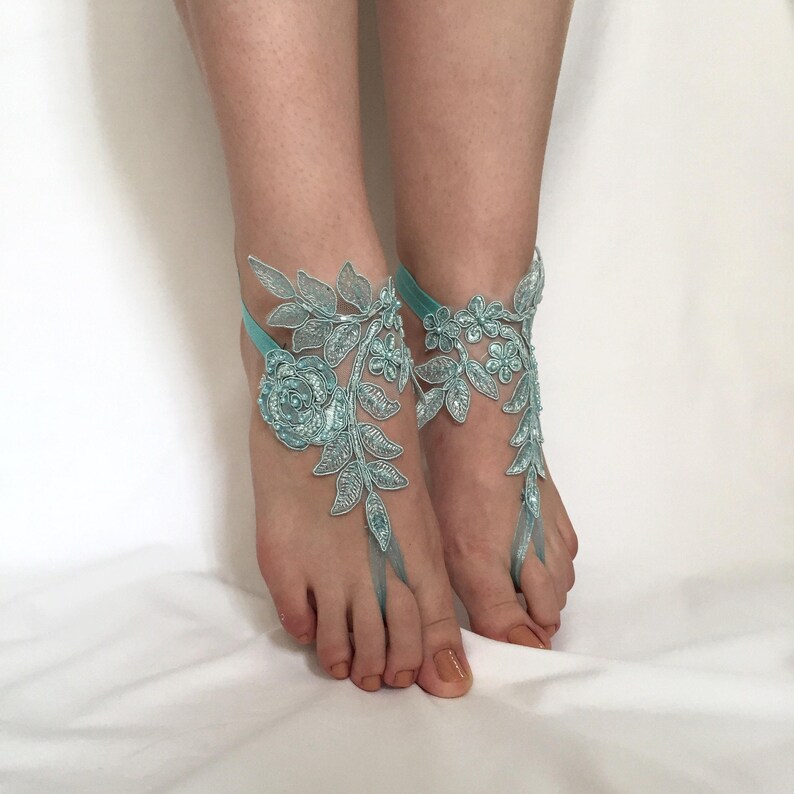 May include: A pair of light blue barefoot sandals featuring intricate floral and leaf lace embroidery. The design includes a central rose motif with delicate vine patterns that extend across the top of the foot, secured by a thin elastic strap.