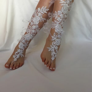 May include: A pair of white lace footless sandals featuring intricate floral embroidery, clear sequins, and small white beads. The design extends from the toe loop up the front of the lower leg, secured with thin white ribbons tied around the ankles.