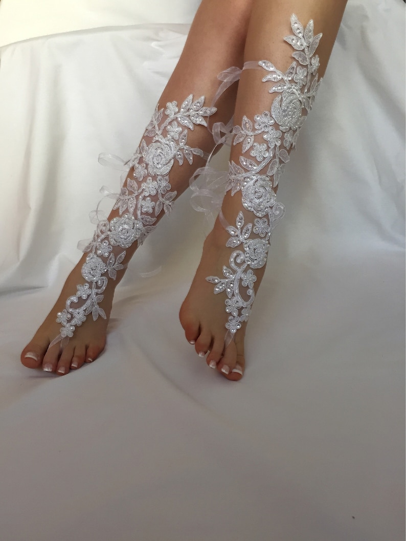 May include: A pair of white lace footless sandals featuring intricate floral embroidery, clear sequins, and small white beads. The design extends from the toe loop up the front of the lower leg, secured with thin white ribbons tied around the ankles.
