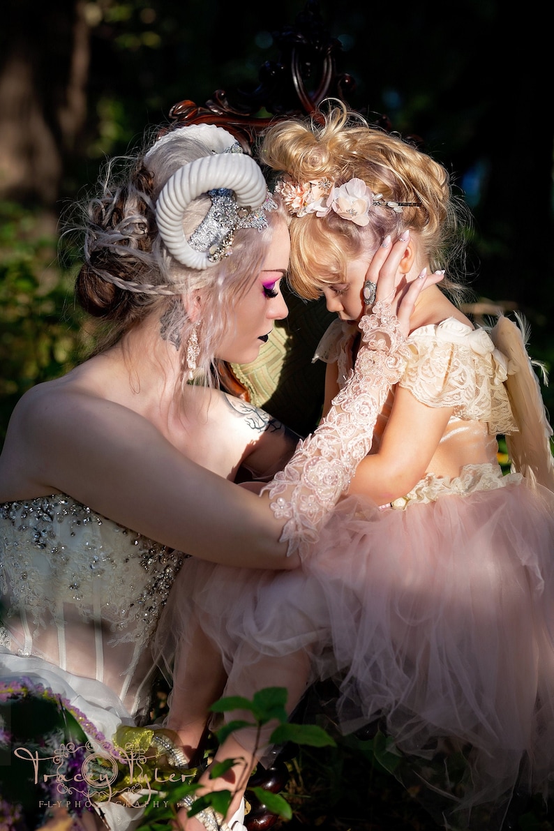 May include: Two people, one wearing a white dress with a silver and white horned headpiece and the other wearing a pink tulle dress with a white lace bodice and a white lace glove, are sitting together and looking at each other. They are both smiling.
