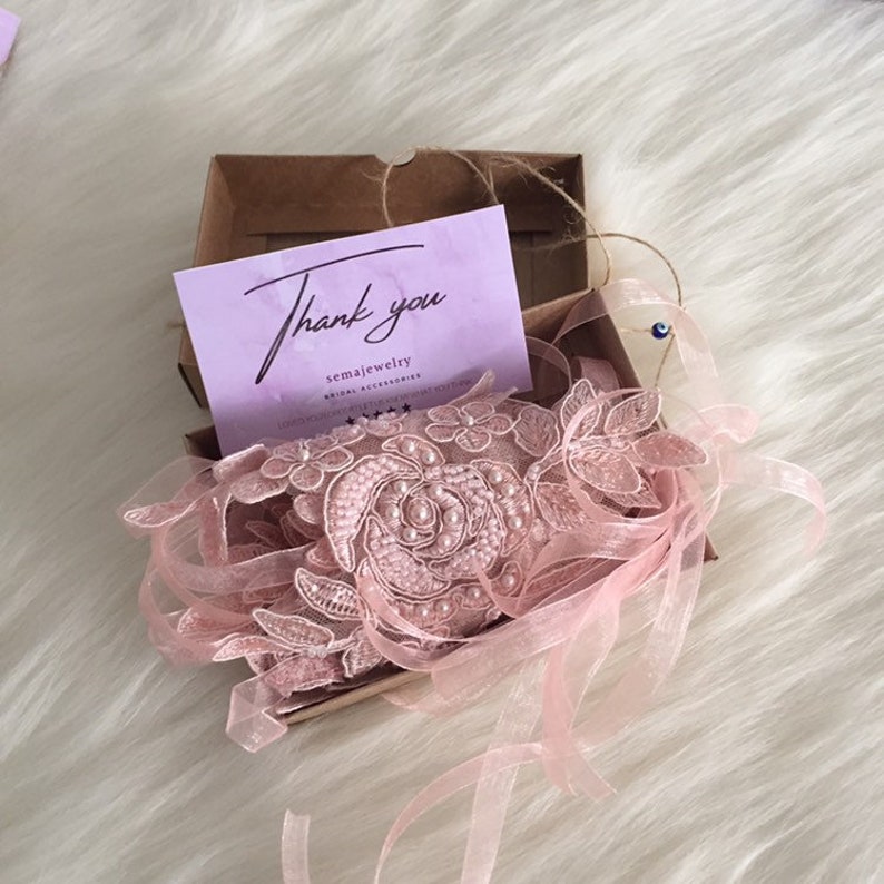 May include: A light pink lace garter featuring floral embroidery and pearl accents, packaged in a brown cardboard box with a light purple card that reads Thank you semajewelry. The garter includes sheer ribbon ties and a small blue evil eye charm.