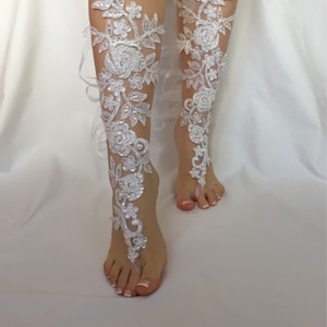 May include: A pair of white lace barefoot sandals featuring intricate floral embroidery, clear sequins, and small beads. The design extends from the toes up the front of the lower leg, secured with delicate white ribbons tied around the ankles.