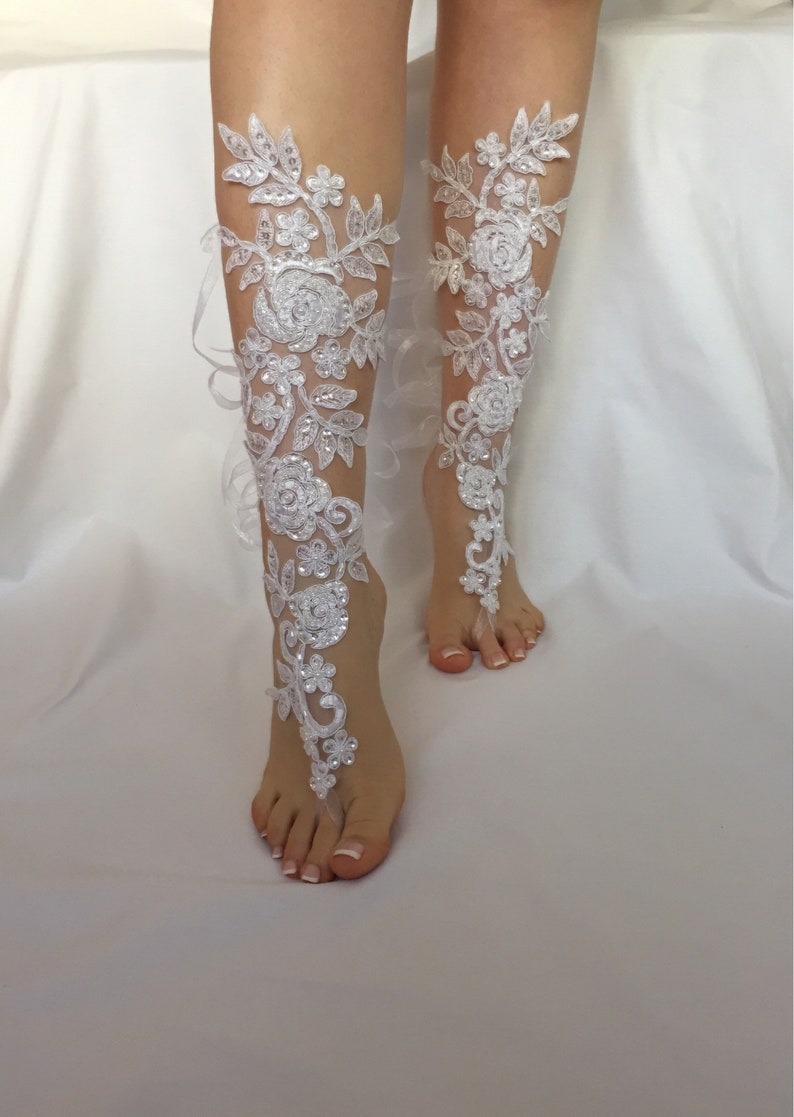 May include: A pair of white lace barefoot sandals featuring intricate floral embroidery, clear sequins, and small beads. The design extends from the toes up the front of the lower leg, secured with delicate white ribbons tied around the ankles.