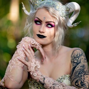 May include: A woman with white hair and white contact lenses wears a white and silver crown with horns. She has pink eyeshadow and black lipstick. She is wearing a white lace dress and has a large tattoo on her arm.
