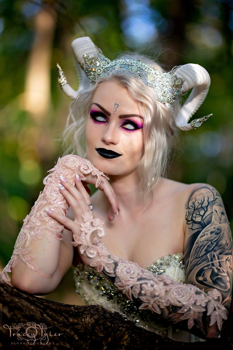 May include: A woman with white hair and white contact lenses wears a white and silver crown with horns. She has pink eyeshadow and black lipstick. She is wearing a white lace dress and has a large tattoo on her arm.