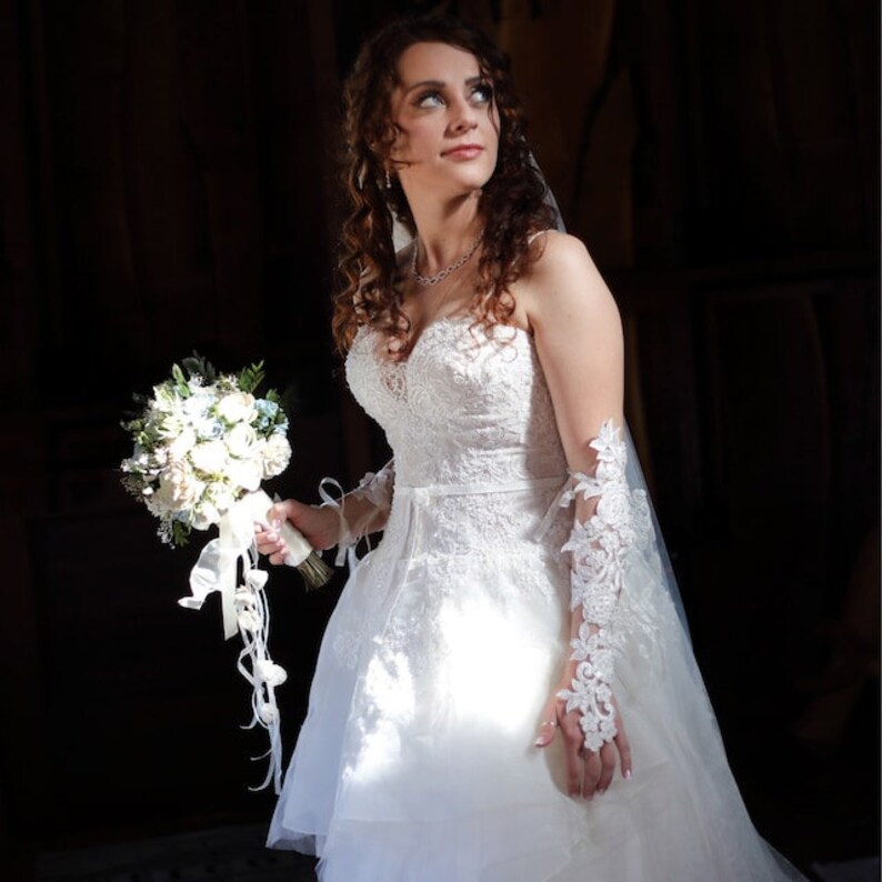 May include: A bride in a white lace wedding dress with a long veil, holding a bouquet of white and blue flowers. She is looking up and smiling.
