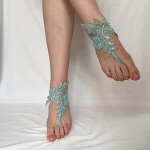May include: A pair of light blue lace barefoot sandals featuring intricate floral and leaf embroidery with delicate bead embellishments. The design wraps around the ankle and includes a loop that secures around the second toe, creating a decorative accessory for the feet.
