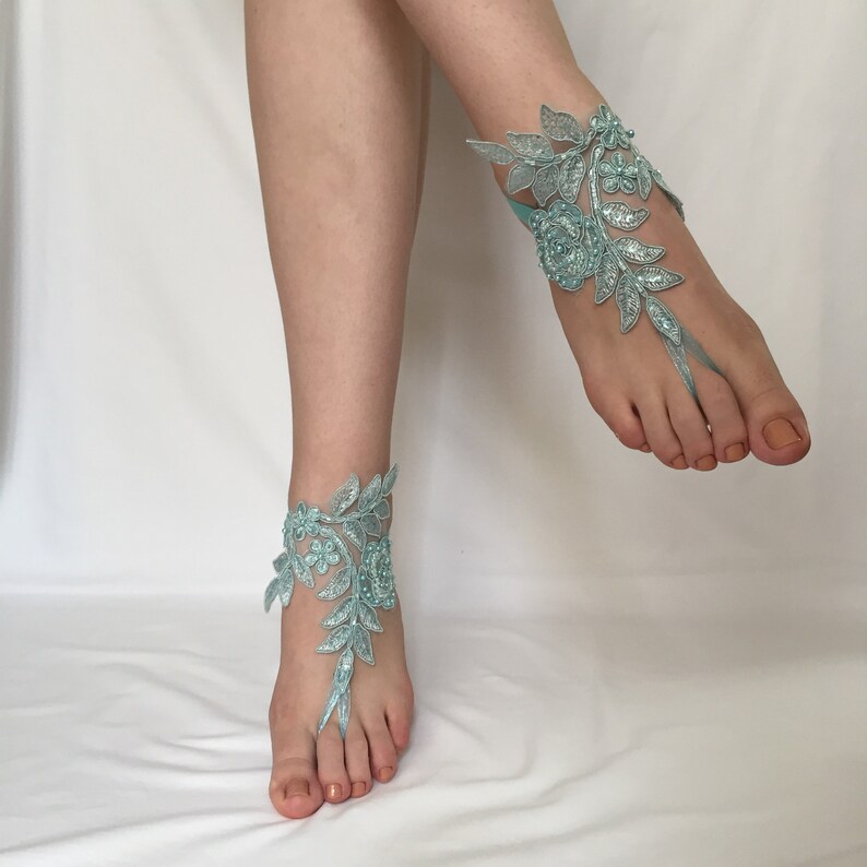 May include: A pair of light blue lace barefoot sandals featuring intricate floral and leaf embroidery with delicate bead embellishments. The design wraps around the ankle and includes a loop that secures around the second toe, creating a decorative accessory for the feet.