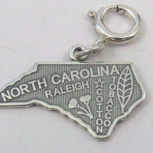 May include: Silver charm shaped like the state of North Carolina. The charm features the state name, the city of Raleigh, and the words "Cotton, Tobacco".