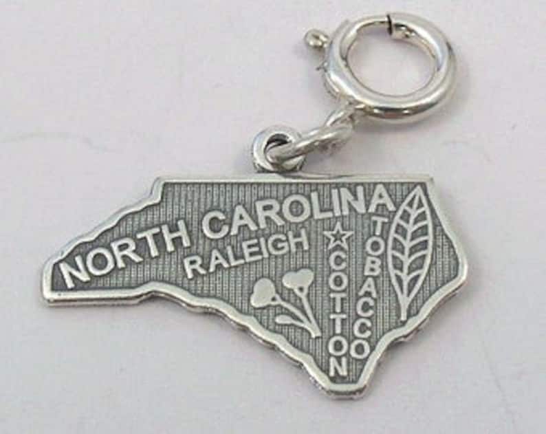 May include: Silver charm shaped like the state of North Carolina. The charm features the state name, the city of Raleigh, and the words "Cotton, Tobacco".