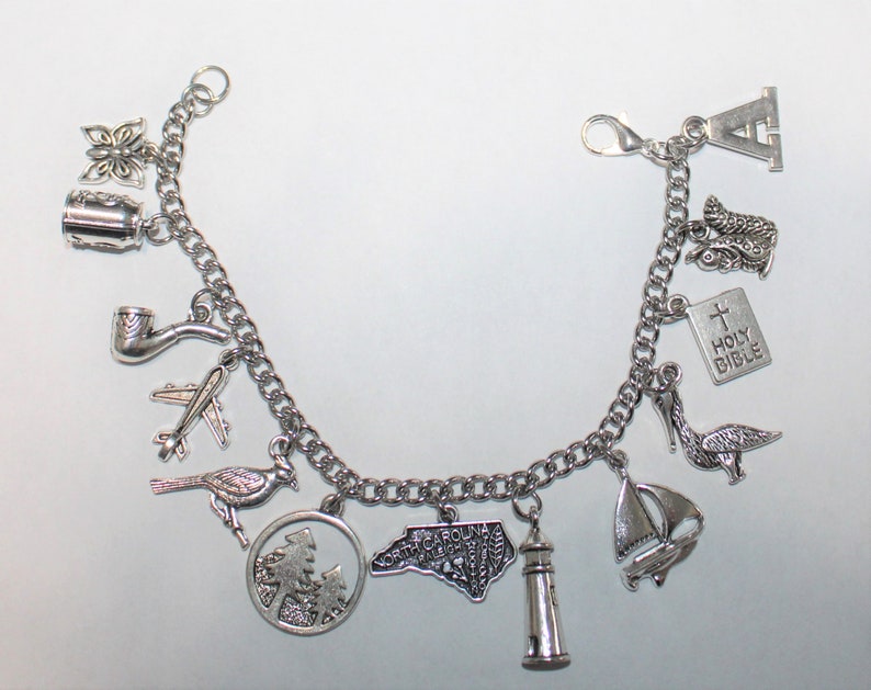 May include: A silver charm bracelet featuring a variety of charms, including a butterfly, a pipe, an aeroplane, a bird, a circle of trees, a state of North Carolina, a lighthouse, a sailboat, a pelican, a boot, a book with the words "Holy Bible", and the letter "A".