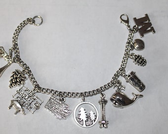 May include: Silver charm bracelet featuring various charms, including a whale, Space Needle, coffee cup, and a charm that says "Public Market Center". The bracelet is made of a chain link design.