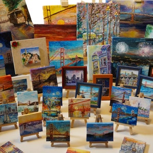 Puede incluir: Una colecci&oacute;n de peque&ntilde;as pinturas enmarcadas en caballetes, que representan diversas escenas del Puente Golden Gate en San Francisco. Las pinturas son de diversos estilos y colores, y algunas incluyen fuegos artificiales, paisajes urbanos y paisajes.
