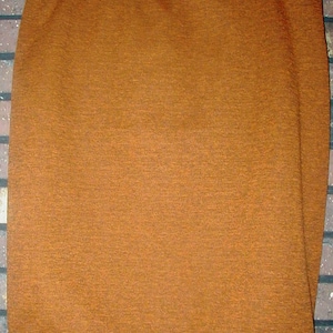 May include: A solid, knee-length skirt in a warm, earthy brown hue. The skirt is made of a textured fabric and hangs from a simple waistband. It is displayed on a hanger against a brick wall.