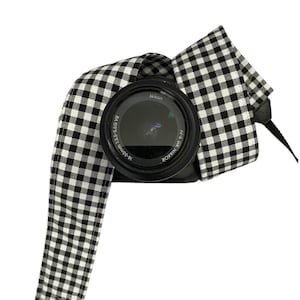 Pode incluir: Uma correia de câmera xadrez em preto e branco com uma lente de câmera preta que diz "NIKKOR DX AF-S 18-55mm 1:3.5-5.6G VR" e "Nikon".