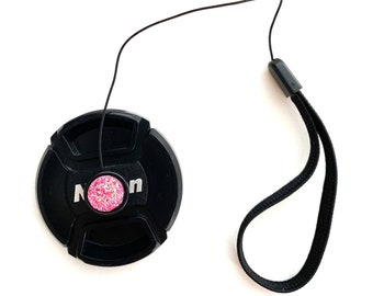 May include: Black camera lens cap with a pink sparkly button and a black strap.