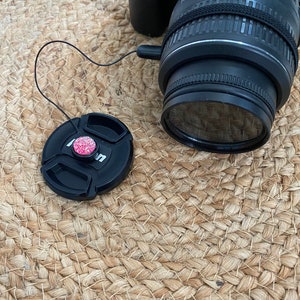 May include: A black camera lens with a black lens cap attached to it by a string. The lens cap has a pink and white decorative button.