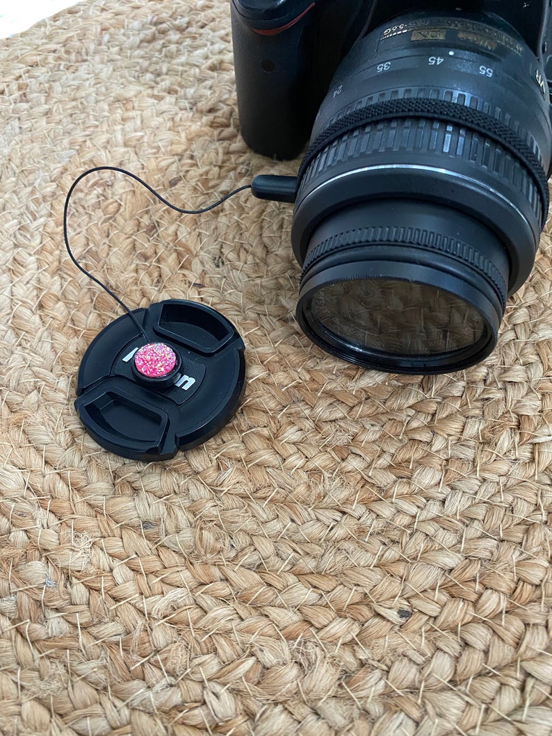 May include: A black camera lens with a black lens cap attached to it by a string. The lens cap has a pink and white decorative button.