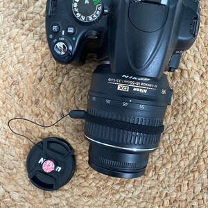 May include: A black Nikon DSLR camera with a black lens attached. The camera has a variety of buttons and dials, including a scene mode dial, a zoom ring, and a focus ring. The lens has a silver band around it and the words "Nikon DX AF-S NIKKOR 18-55mm 1:3.5-5.6G VR" printed on it.