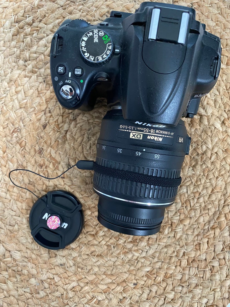 May include: A black Nikon DSLR camera with a black lens attached. The camera has a variety of buttons and dials, including a scene mode dial, a zoom ring, and a focus ring. The lens has a silver band around it and the words "Nikon DX AF-S NIKKOR 18-55mm 1:3.5-5.6G VR" printed on it.