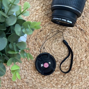 May include: A black camera lens cap with a pink and white glitter design and the word "NOON" on it. The lens cap is attached to a black string.