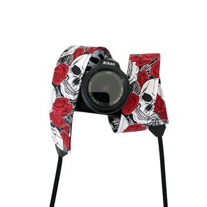 May include: A black camera with a white and red skull and rose print camera strap. The strap has the word "Nikon" printed on it.