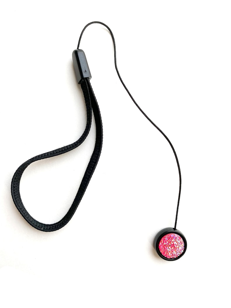 May include: A black camera strap with a pink glitter button. The strap is made of a soft, flexible material and has a simple design.