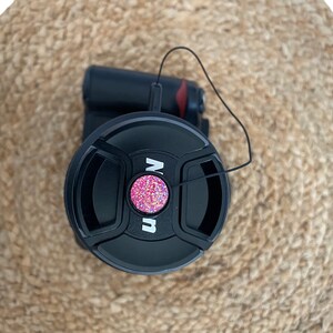 May include: Black camera lens cap with a pink glitter button and a black string attached.