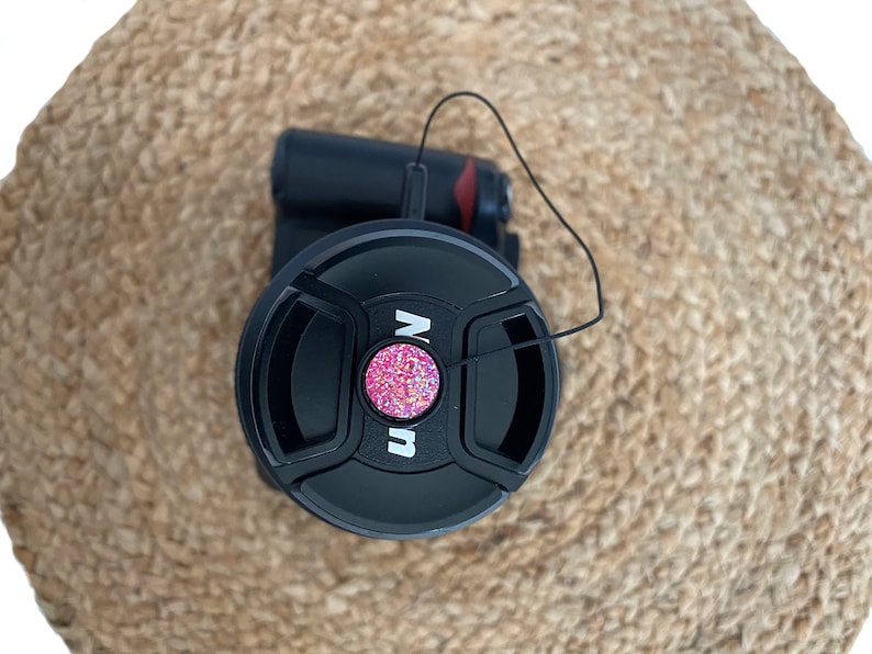 May include: Black camera lens cap with a pink glitter button and a black string attached.