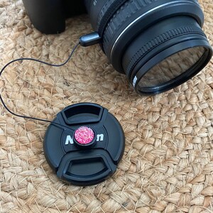 May include: A black camera lens cap with a pink sparkly button. The cap is attached to a black string.