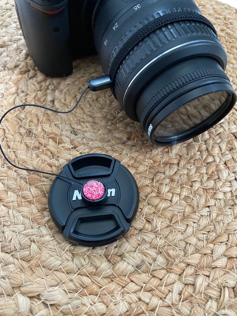 May include: A black camera lens cap with a pink sparkly button. The cap is attached to a black string.