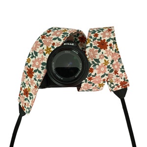May include: A black camera with a floral patterned camera strap. The strap is pink, white and green with red flowers.