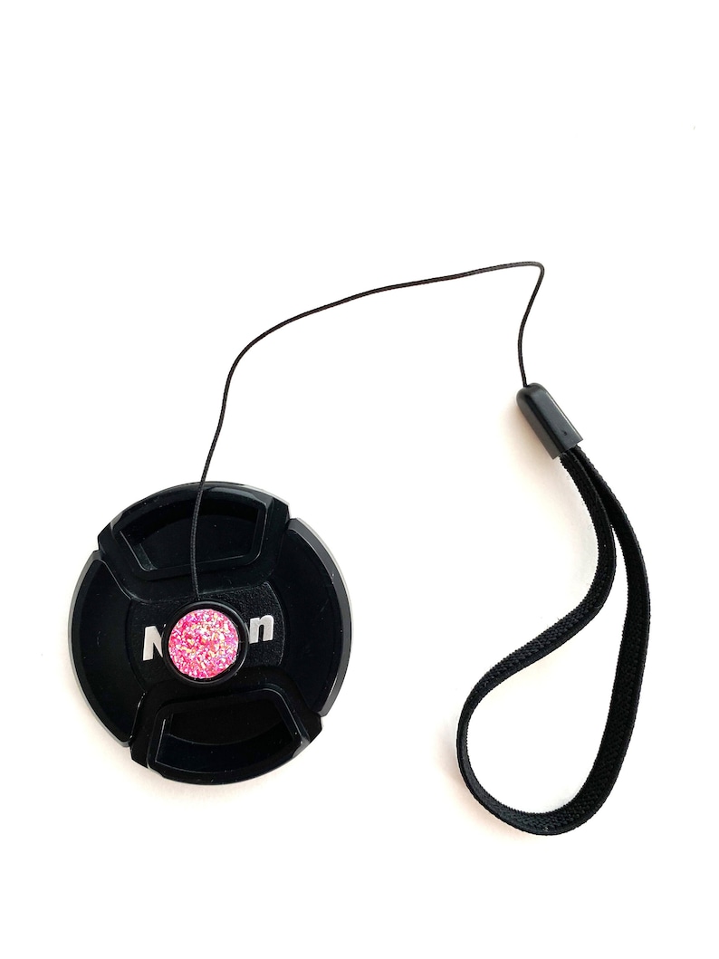 May include: Black camera lens cap with a pink sparkly button and a black strap.
