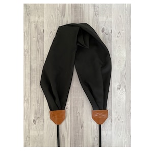 Pode incluir: Uma alça de câmera preta com extremidades de couro marrom e fixações de cordão de nylon preto. A alça é feita de um tecido macio e drapeado. As extremidades de couro têm um design costurado. O fundo é um grão de madeira cinza claro.